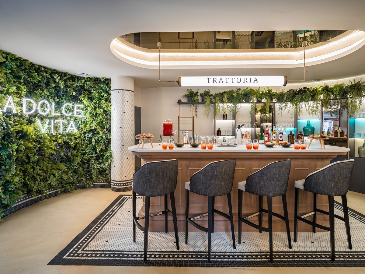 A curved wooden bar with four upholstered stools on a mosaic floor, bottles and cocktails along the counter, a neon La Dolce Vita sign on a living green wall to one side and a lit Trattoria sign above.