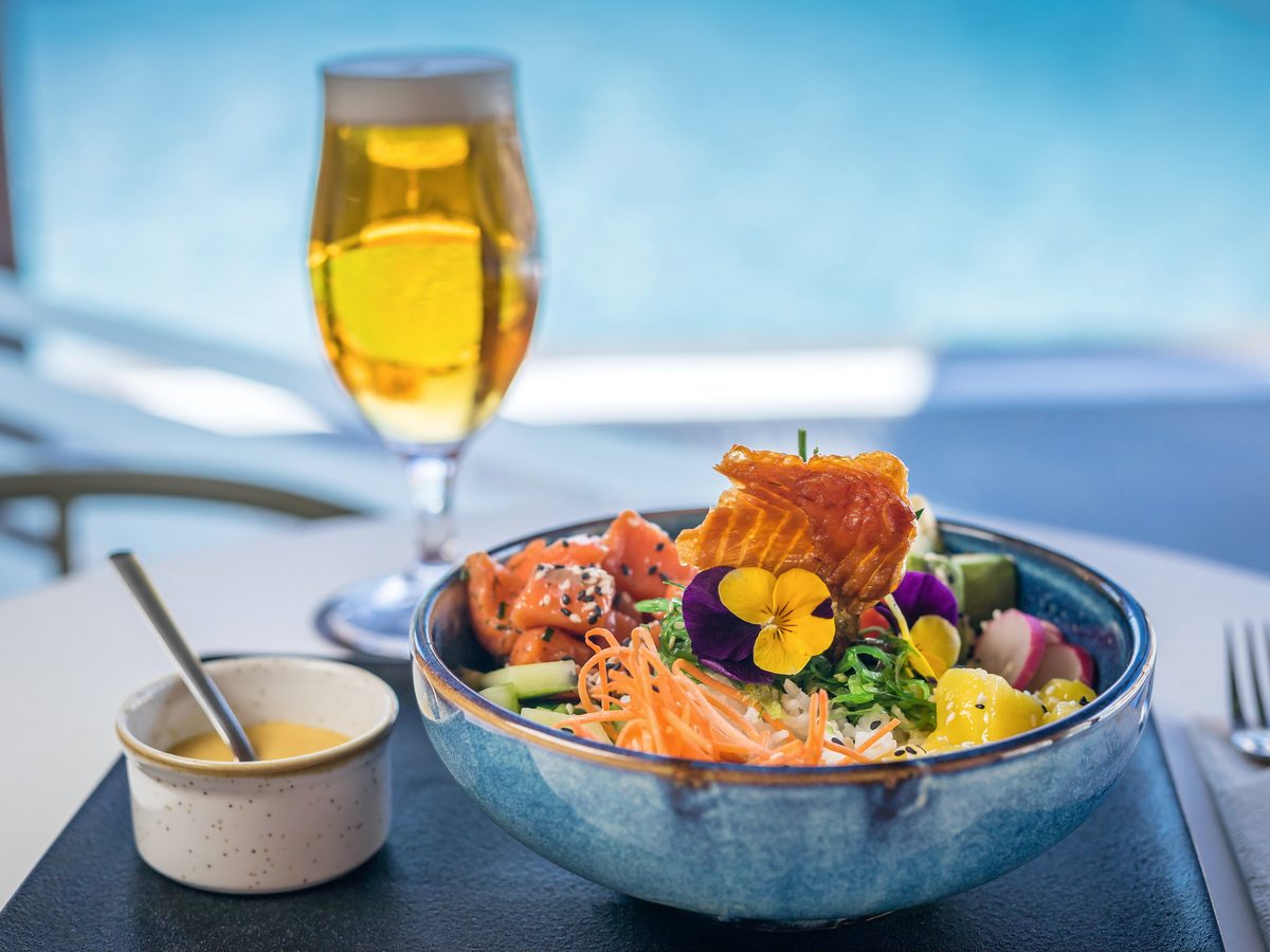 A blue bowl of salmon poke with rice, shredded carrot, mango, radish and edible flowers, with a sauce pot and a glass of beer, against a bright blue pool.