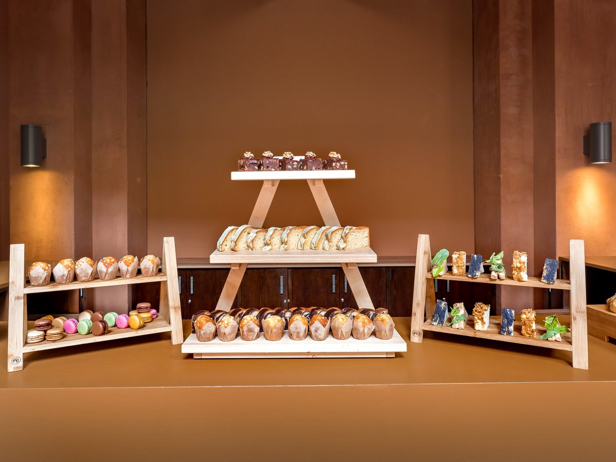 A tiered wooden stand of cakes, muffins, macarons and small desserts arranged on a counter against a warm brown wall.
