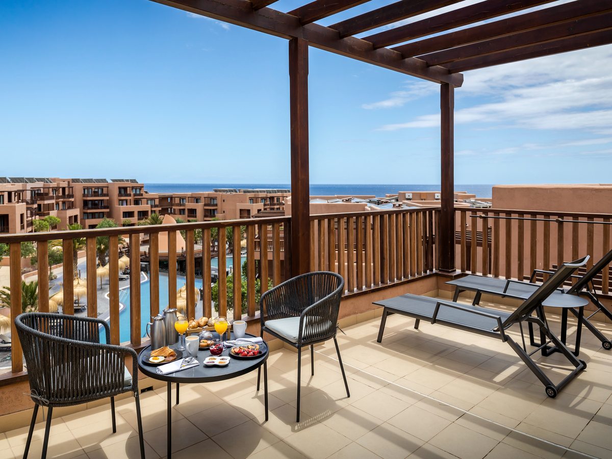 A tiled balcony under a dark wooden pergola with two woven chairs at a round table set with breakfast, a sun lounger to one side, and the resort's pools and the sea beyond the railing.