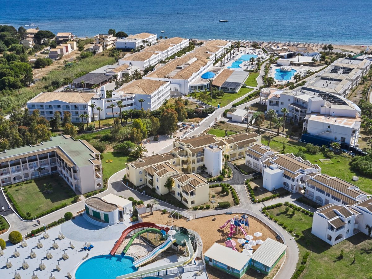 An aerial view of the resort's white low-rise blocks and green gardens, with pools, coloured water slides and a playground below, and the beach and blue sea along the top.