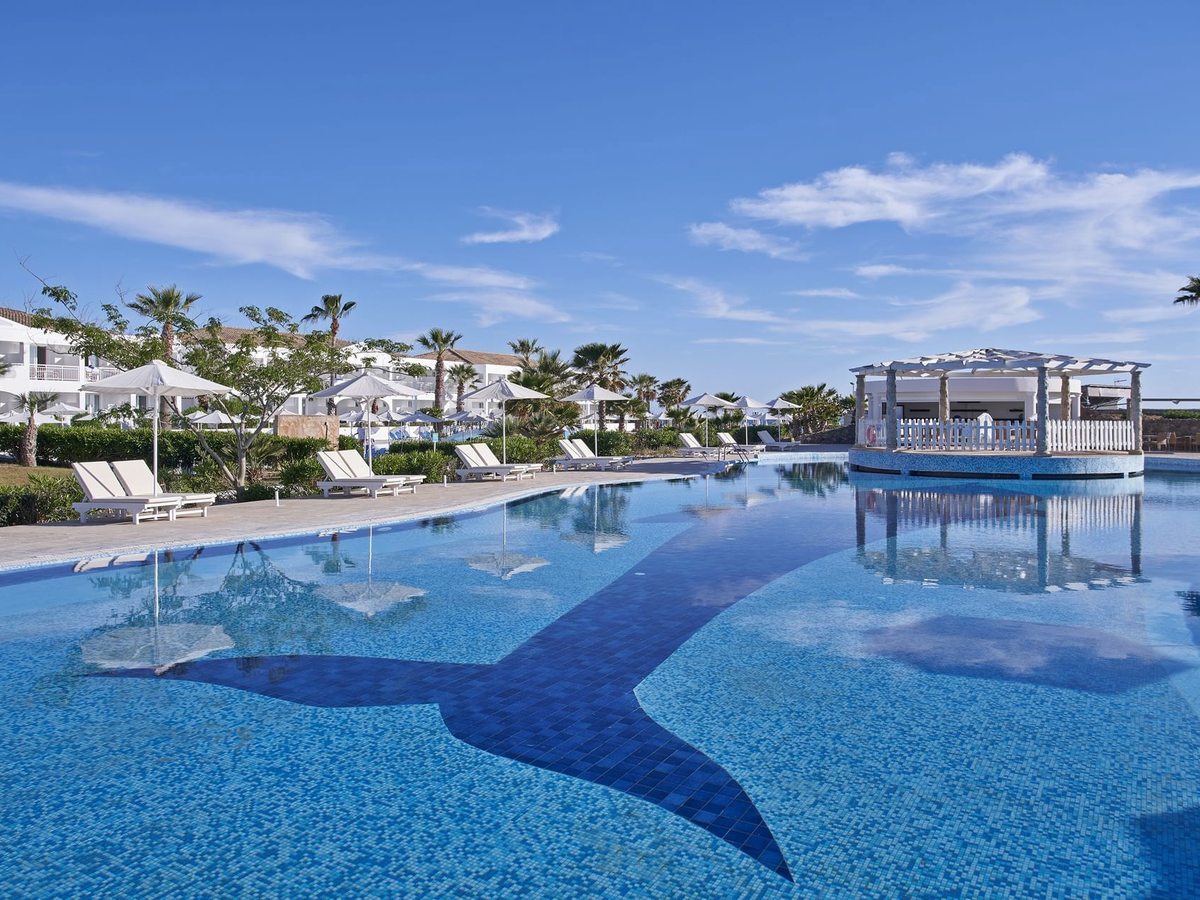 A large blue pool with a dark mosaic tail design across its floor, white loungers and parasols along the edge, palms and low white buildings behind and a covered bar on an island in the water.