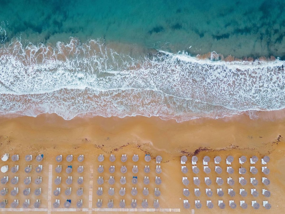 An aerial view looking straight down on rows of thatched parasols and sunbeds on golden sand, with waves breaking along the shore and turquoise water beyond.