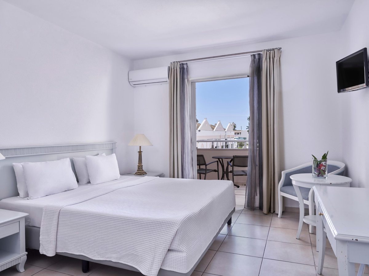A white double room with a bed in white linen, a small table and armchair, a wall-mounted television and glass doors open to a balcony with a table and two chairs.
