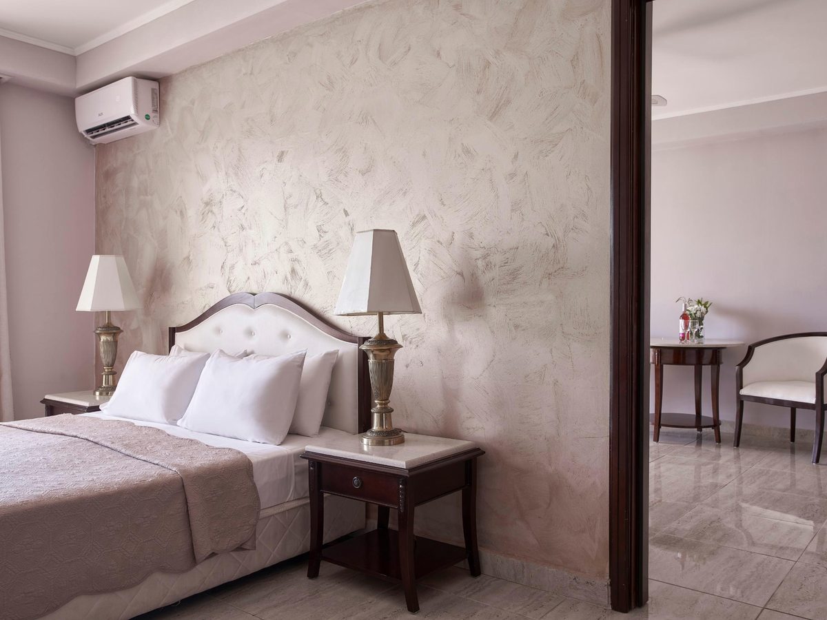 A suite bedroom with a double bed, a buttoned headboard and brass lamps against a textured cream wall, with a doorway through to a sitting area with a round table and armchair.