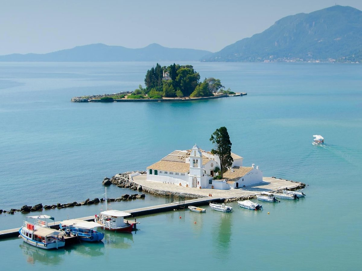 A small white monastery with a bell tower on a low islet linked to the shore by a causeway, fishing boats moored alongside and a wooded islet in calm blue sea beyond.