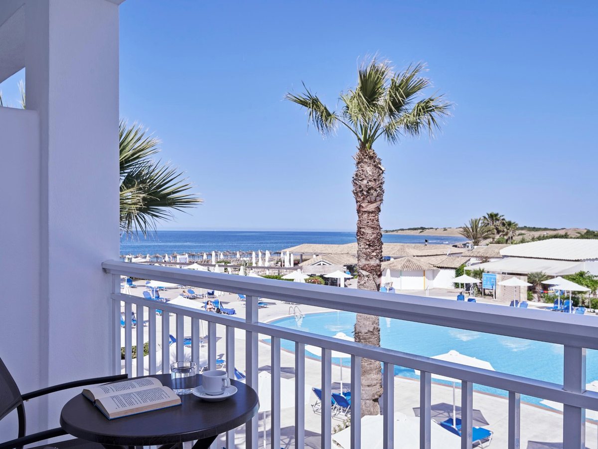 A balcony with a dark table and two chairs, a book and a cup, looking past a palm to the pool below, parasols on the sand and the open sea.