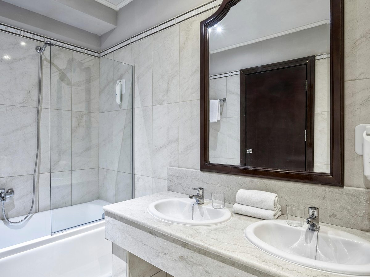 A bathroom with a bath behind a glass screen, twin basins set in a marble counter, a large framed mirror and rolled white towels.