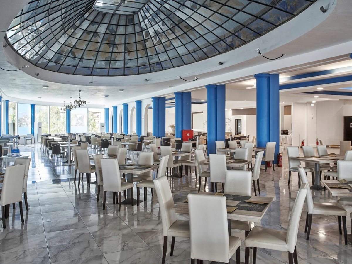 A large restaurant hall with white leather chairs at set tables, blue columns and a domed glass skylight above a marble floor.