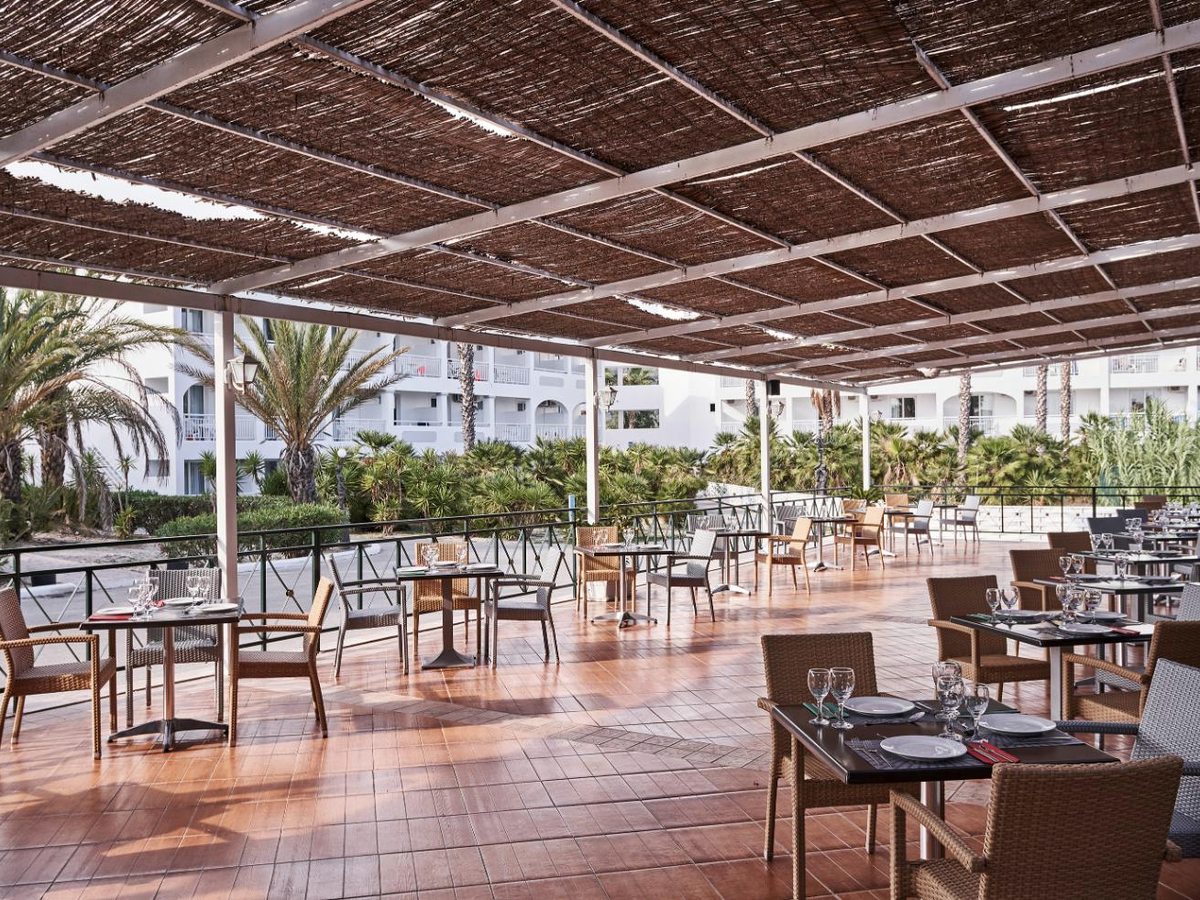A wide open-sided terrace under a reed-covered pergola, with wicker chairs at dark tables on a terracotta floor and white accommodation blocks and palms beyond.