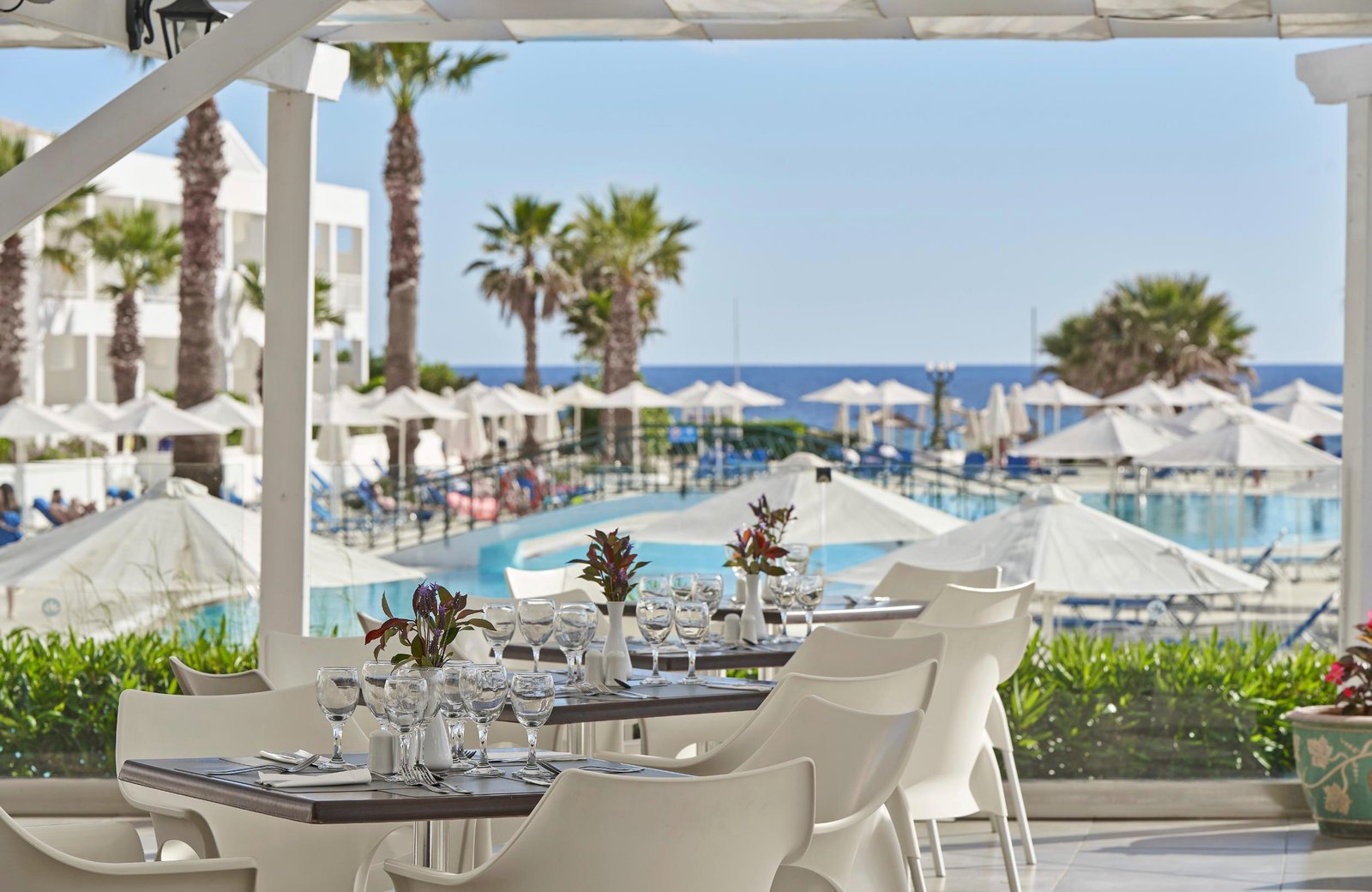 A shaded restaurant terrace with laid tables at Sandy Beach Resort, above a large pool with white parasols, palms and the sea beyond.