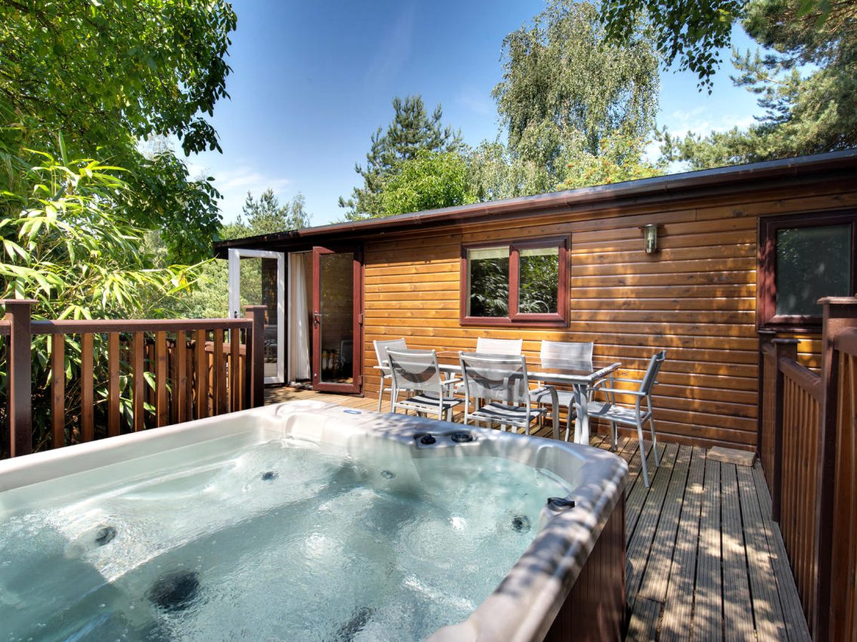 A bubbling hot tub set into a timber deck beside a table and six chairs, outside a lodge with its patio doors open, under a blue sky.