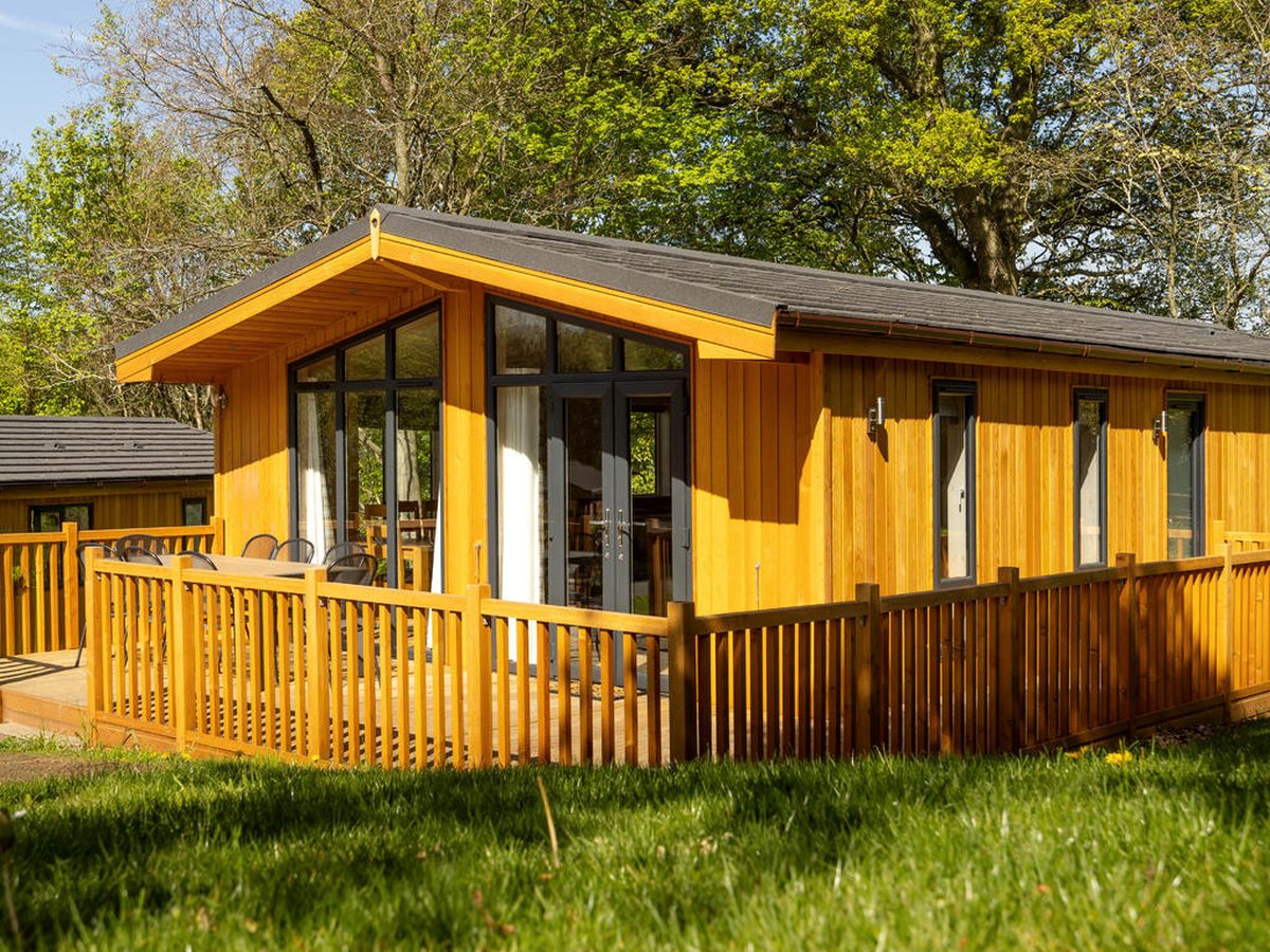 A timber lodge with a pitched roof and full-height windows, its patio doors opening onto a railed deck with a table and chairs, on a grassy plot backed by trees.