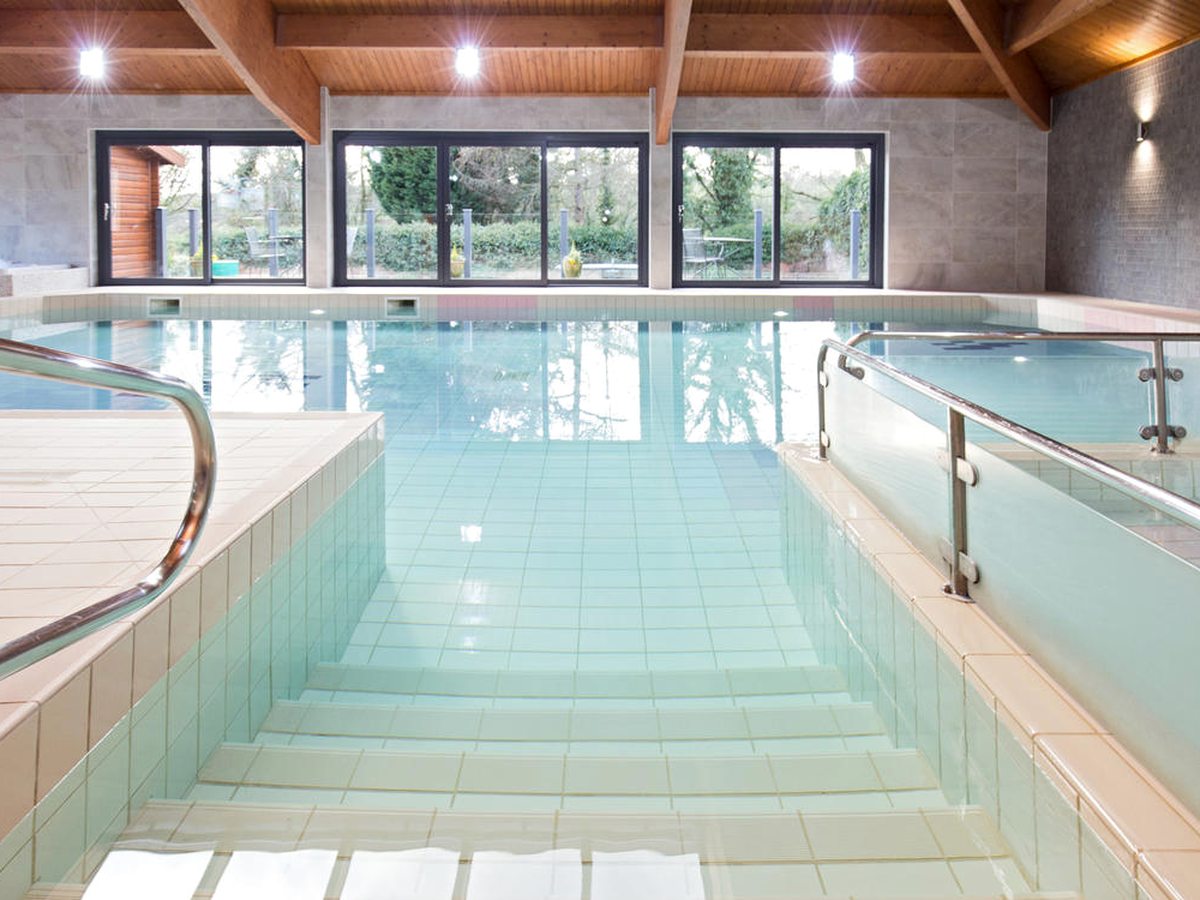 Wide tiled steps leading down into the shallow end of an indoor pool, with metal handrails and a glass-panelled rail alongside.