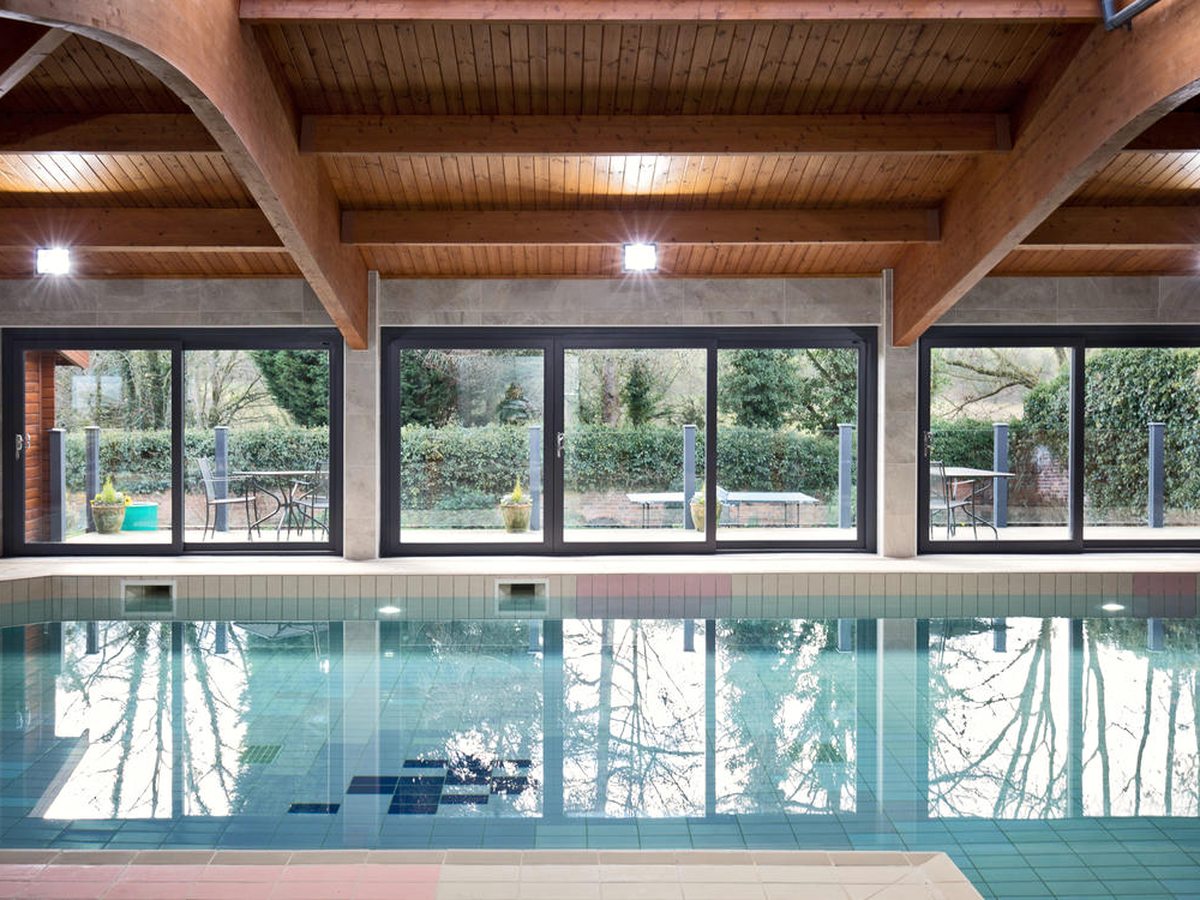 A long indoor swimming pool under a timber-beamed roof, with sliding glass doors along one side onto a hedged terrace.