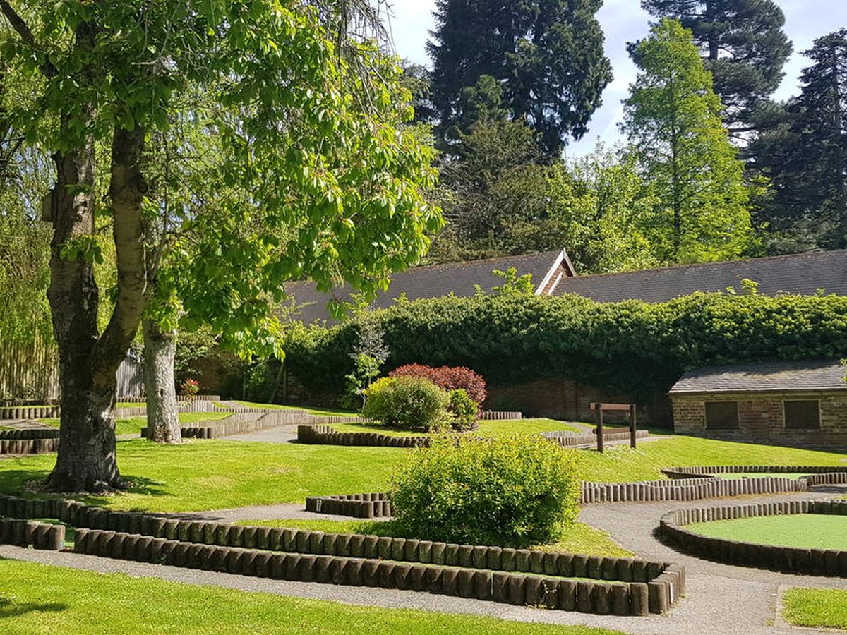 A mini golf course laid out on lawns with log-edged borders and green putting surfaces, among mature trees beside a stone outbuilding.