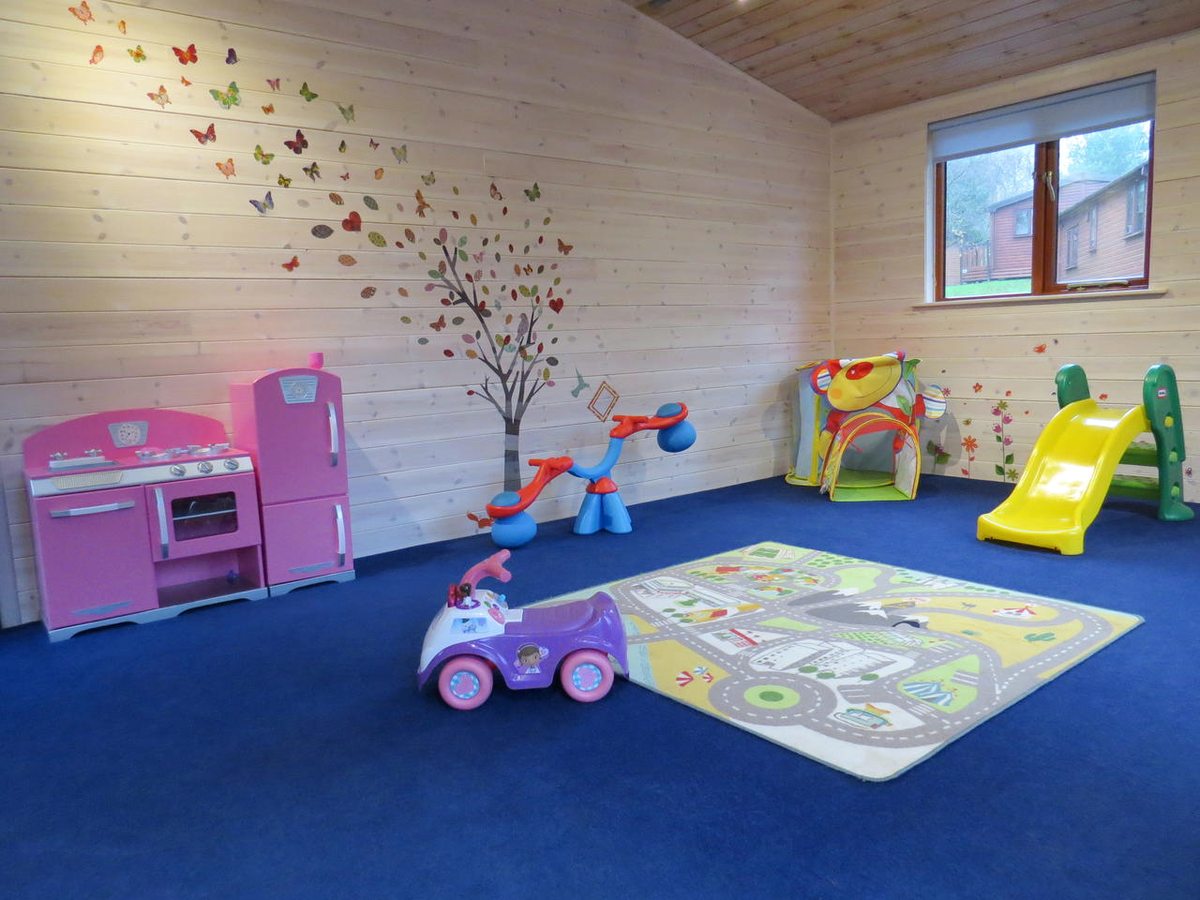 A carpeted playroom in a timber-lined lodge with a pink toy kitchen, ride-on toys, a fabric play tunnel, a small plastic slide and a road-print play mat.