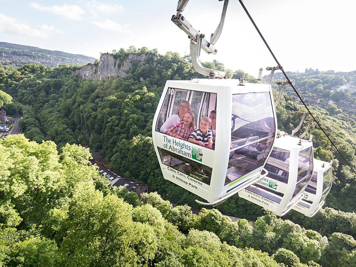Cable cars branded Heights of Abraham on a wire above a wooded valley, with limestone cliffs and a family riding in the nearest one.