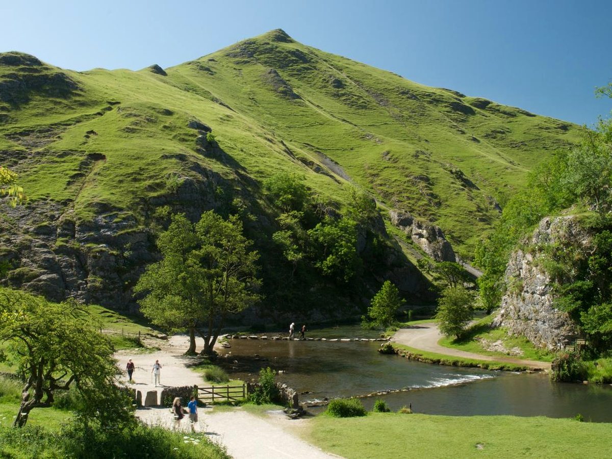 A steep green limestone hill rising above a river valley, with walkers crossing stepping stones at the water's edge.