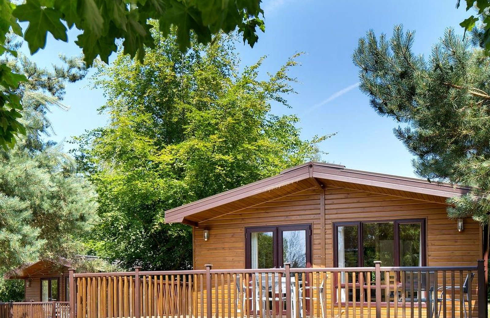 A timber lodge at Sandybrook Lodges with a railed veranda and outdoor seating, among mature trees under a blue sky.