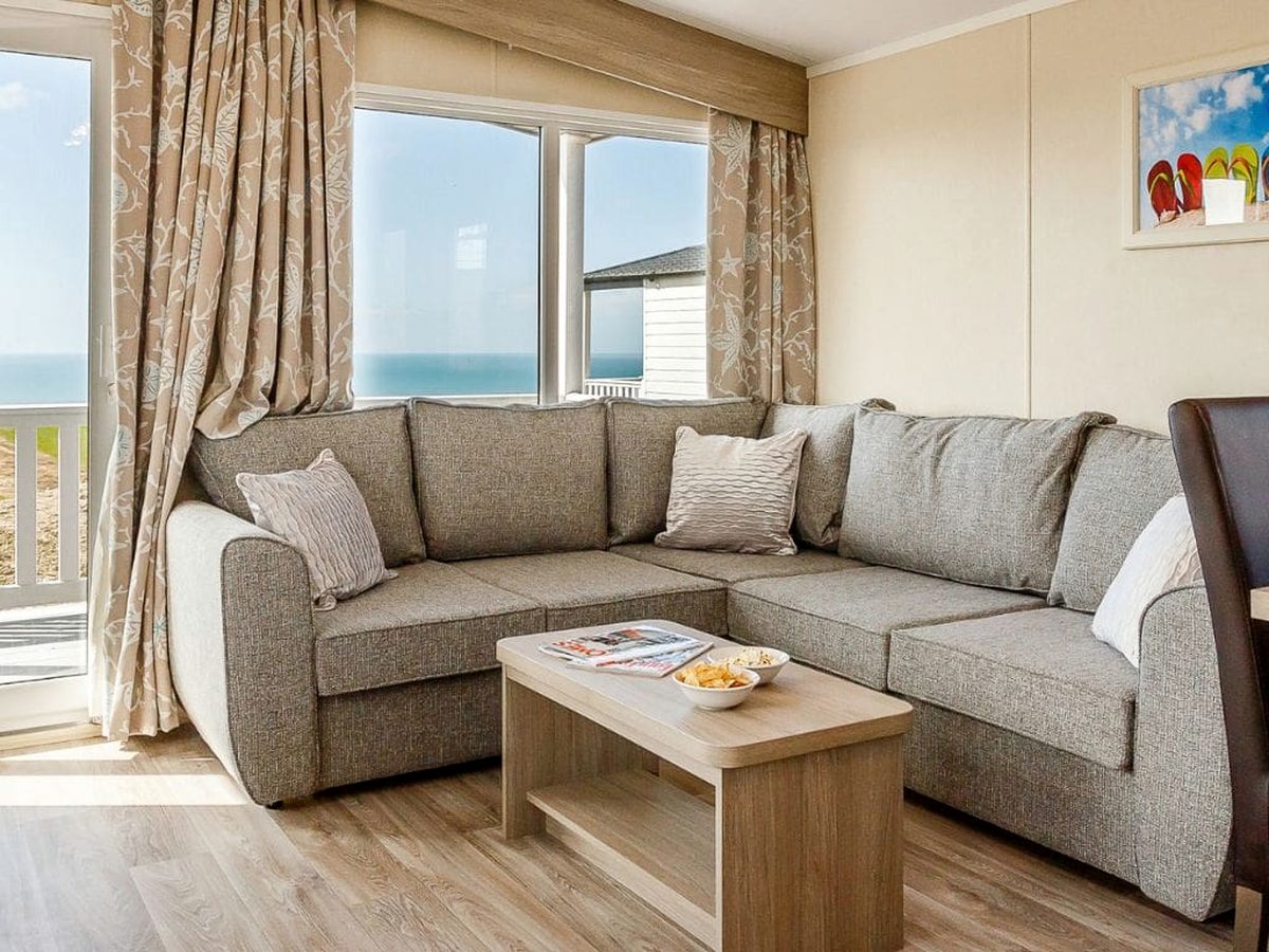 An open-plan lounge with a grey corner sofa, wooden coffee table and armchair, with wide windows out to the sea and patio doors to a deck.