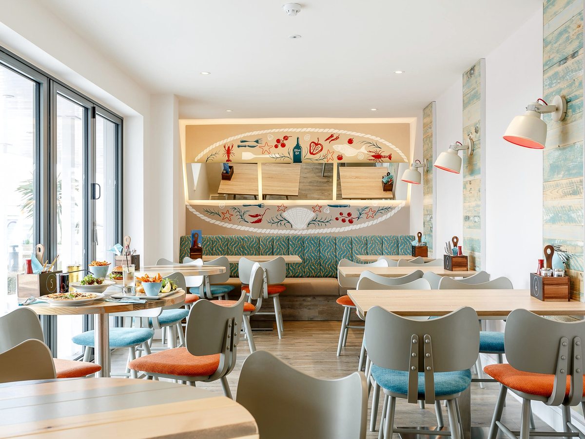 A bright restaurant dining room with pale wooden tables, orange and grey chairs, banquette seating and a painted seaside mural on the far wall.