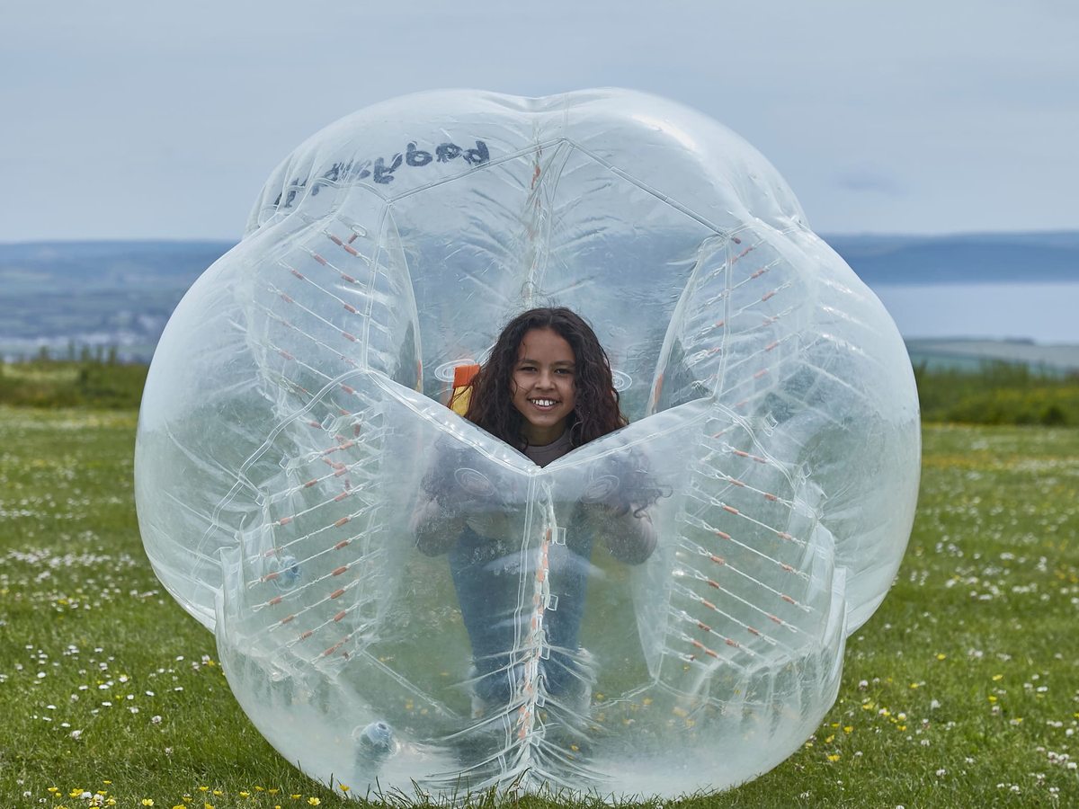 A child inside a large clear inflatable zorb ball on a grassy field, with the coast in the distance.