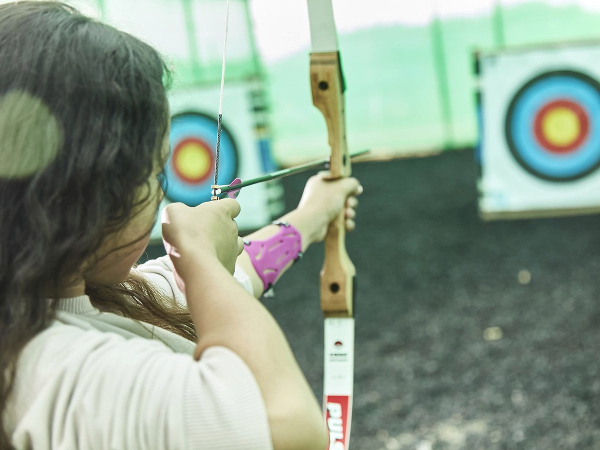 A girl drawing a recurve bow on a netted outdoor archery range, with two round coloured targets on freestanding stands.