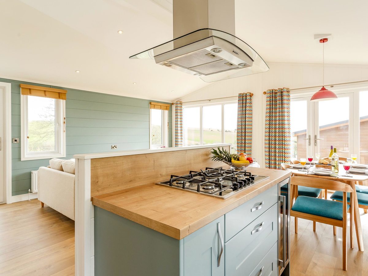 An open-plan kitchen with a wooden-topped island, gas hob and extractor hood, opening onto a dining table with teal and red chairs.