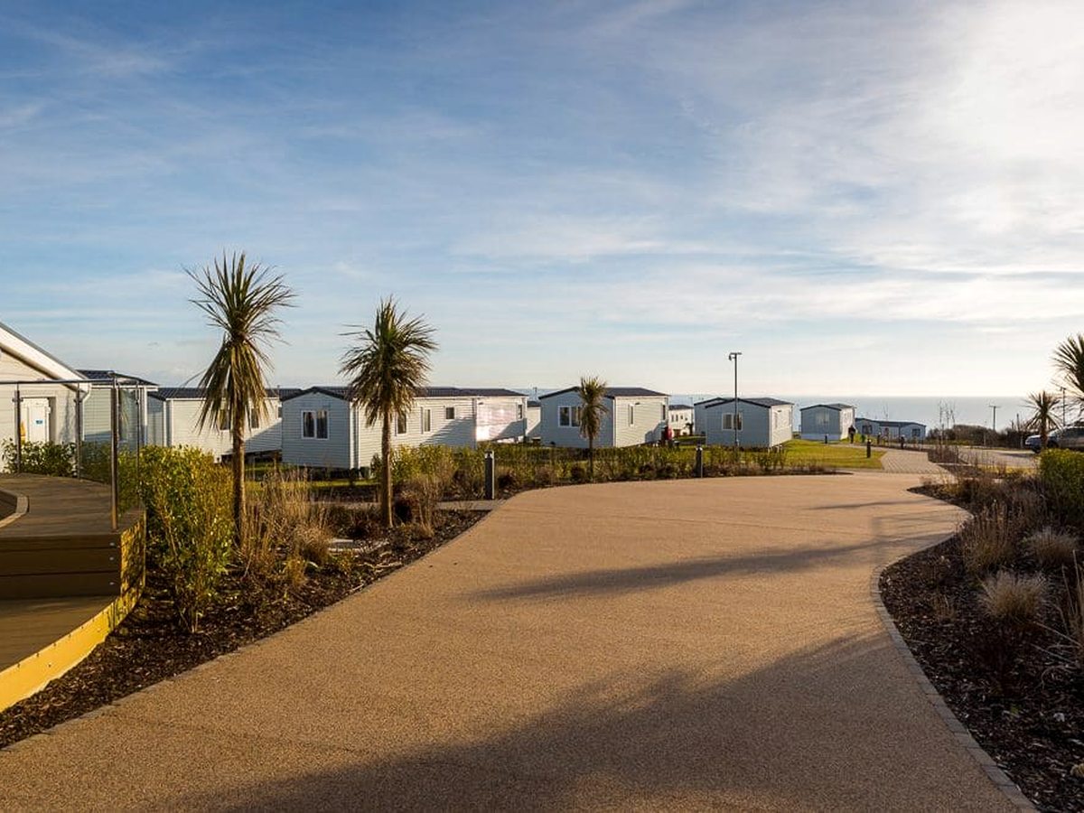 A wide paved road curving between rows of white holiday homes, edged with planting beds and cordyline palms, with the sea in the distance.