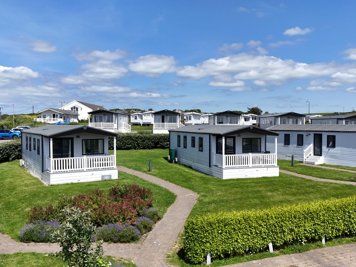 Rows of white holiday homes with railed verandas on mown lawns behind clipped hedges, with a flower bed and paved paths in front, under a blue sky.