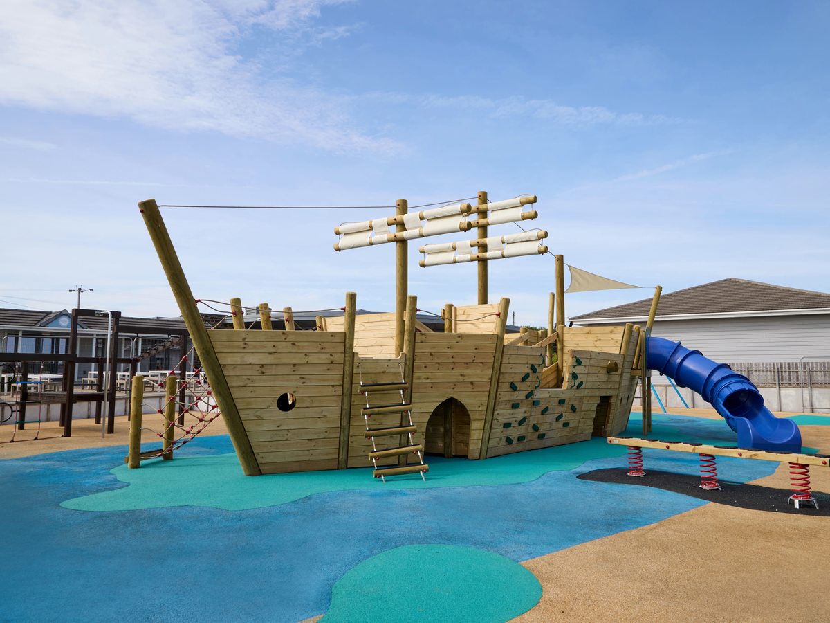 A large wooden pirate-ship play structure with masts, rope rigging, a climbing wall and a blue tube slide, on blue and green safety surfacing.