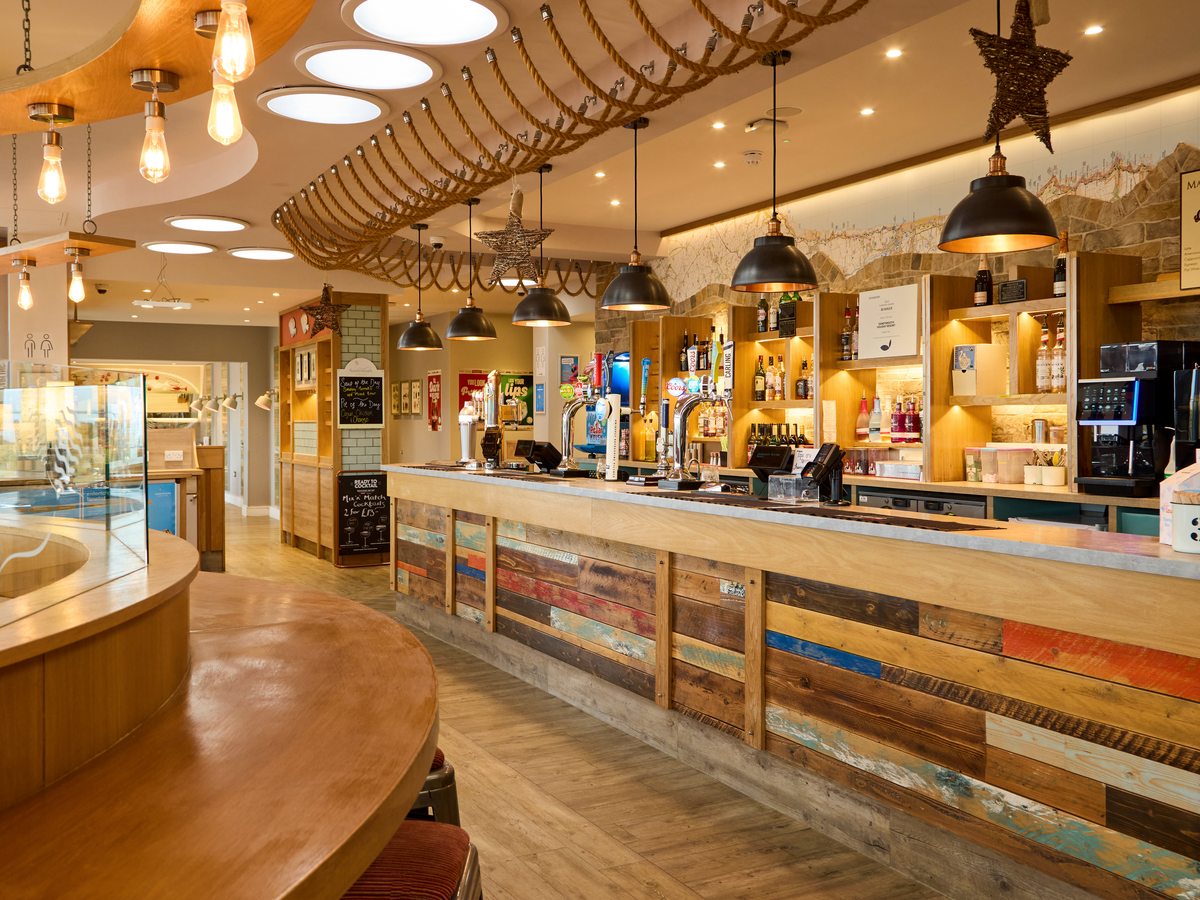 A long bar counter clad in painted reclaimed timber, with draught beer taps, filament pendant lights and shelves of bottles behind.