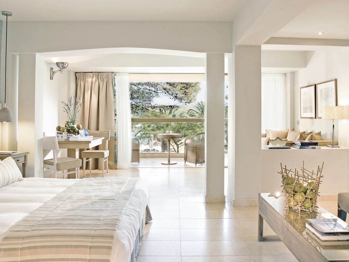 White open-plan room with a double bed, a writing desk and a seating area, and glass doors onto a shaded terrace among pine trees.