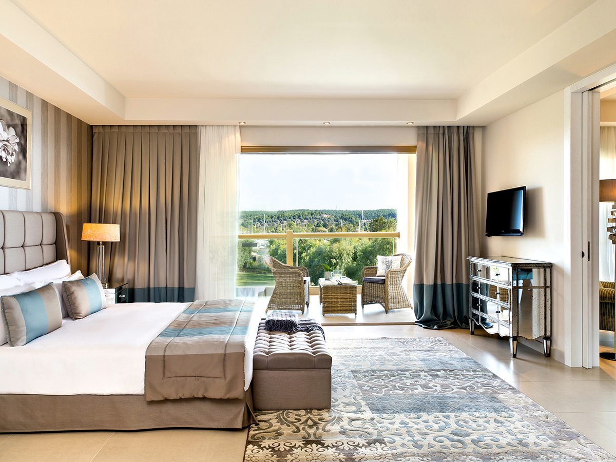 Suite bedroom with a padded headboard, patterned rug and upholstered bench, opening onto a balcony with wicker chairs looking over green treetops.