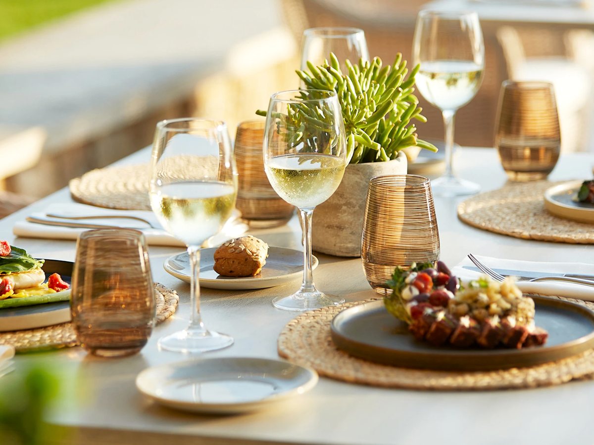 Outdoor dining table set with woven placemats, white wine, amber tumblers, bread and two plated starters in late afternoon light.