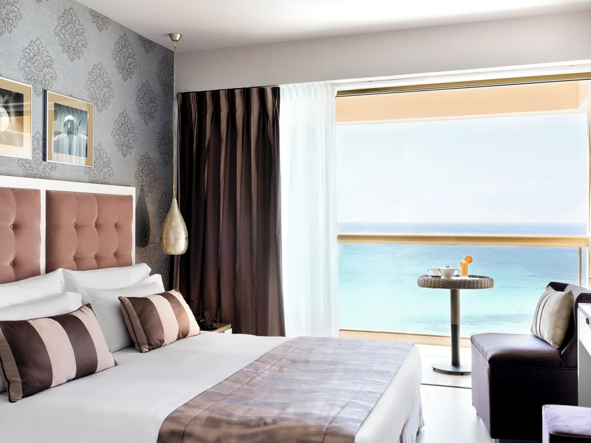 Double room with a pink buttoned headboard, brown and cream striped cushions and patterned wallpaper, with glass doors onto a balcony set with a small table above the sea.