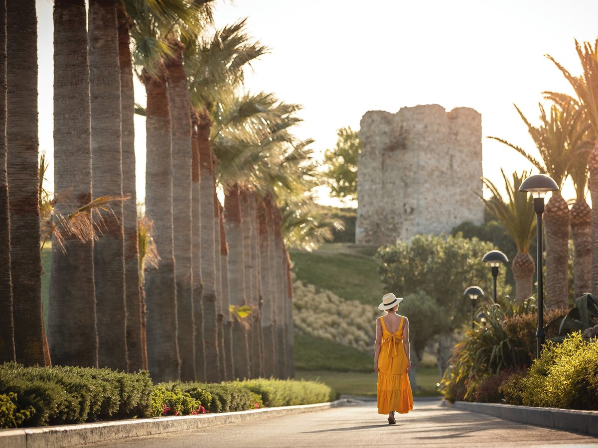 A broad paved path lined with tall palm trees, clipped hedges and flowerbeds, with a ruined stone tower on a grassy rise beyond and a woman in a yellow dress walking ahead.