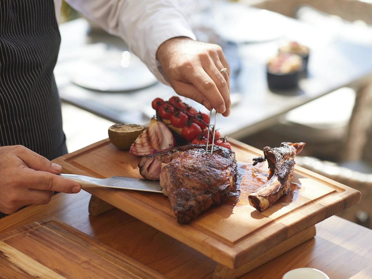 A chef in a striped apron slicing a bone-in grilled beef chop on a wooden board, with charred cherry tomatoes, red onion and lemon alongside.