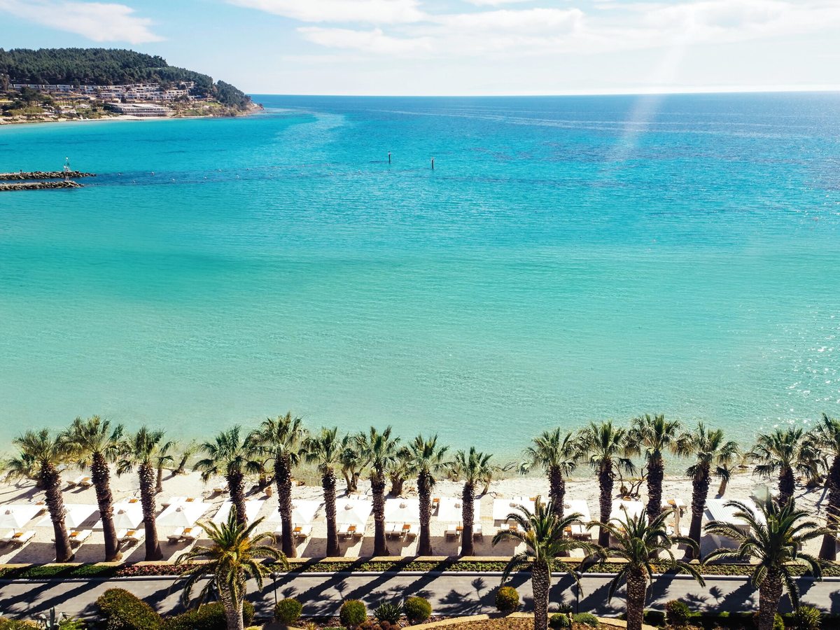 Aerial view of a white-sand beach lined with palms, white parasols and loungers, with clear turquoise shallows, a stone breakwater and a wooded headland beyond.