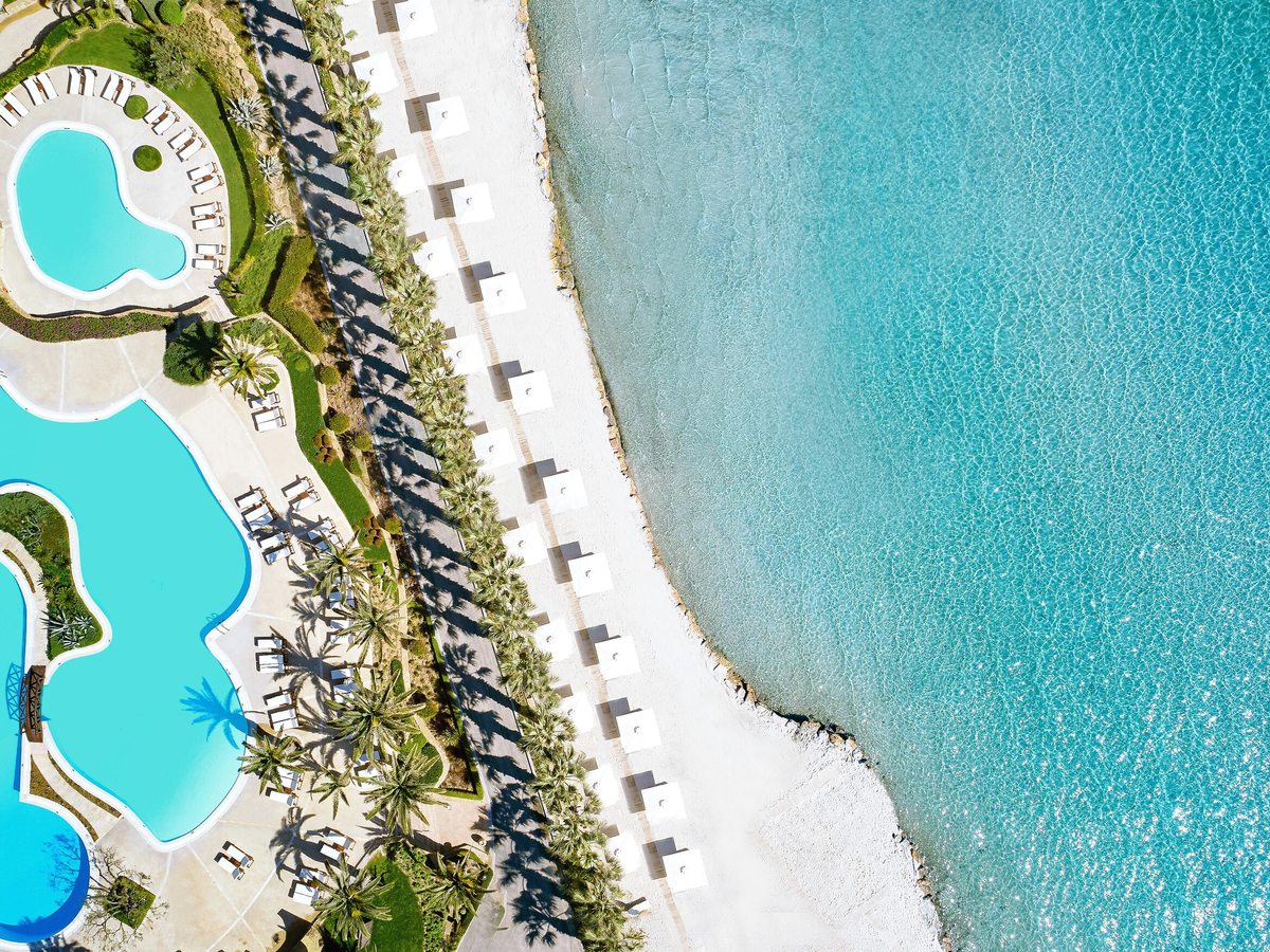 Overhead aerial of two curved pools and their lounger terraces, separated from a white-sand beach by a line of palms and a row of white parasols, with clear turquoise sea alongside.
