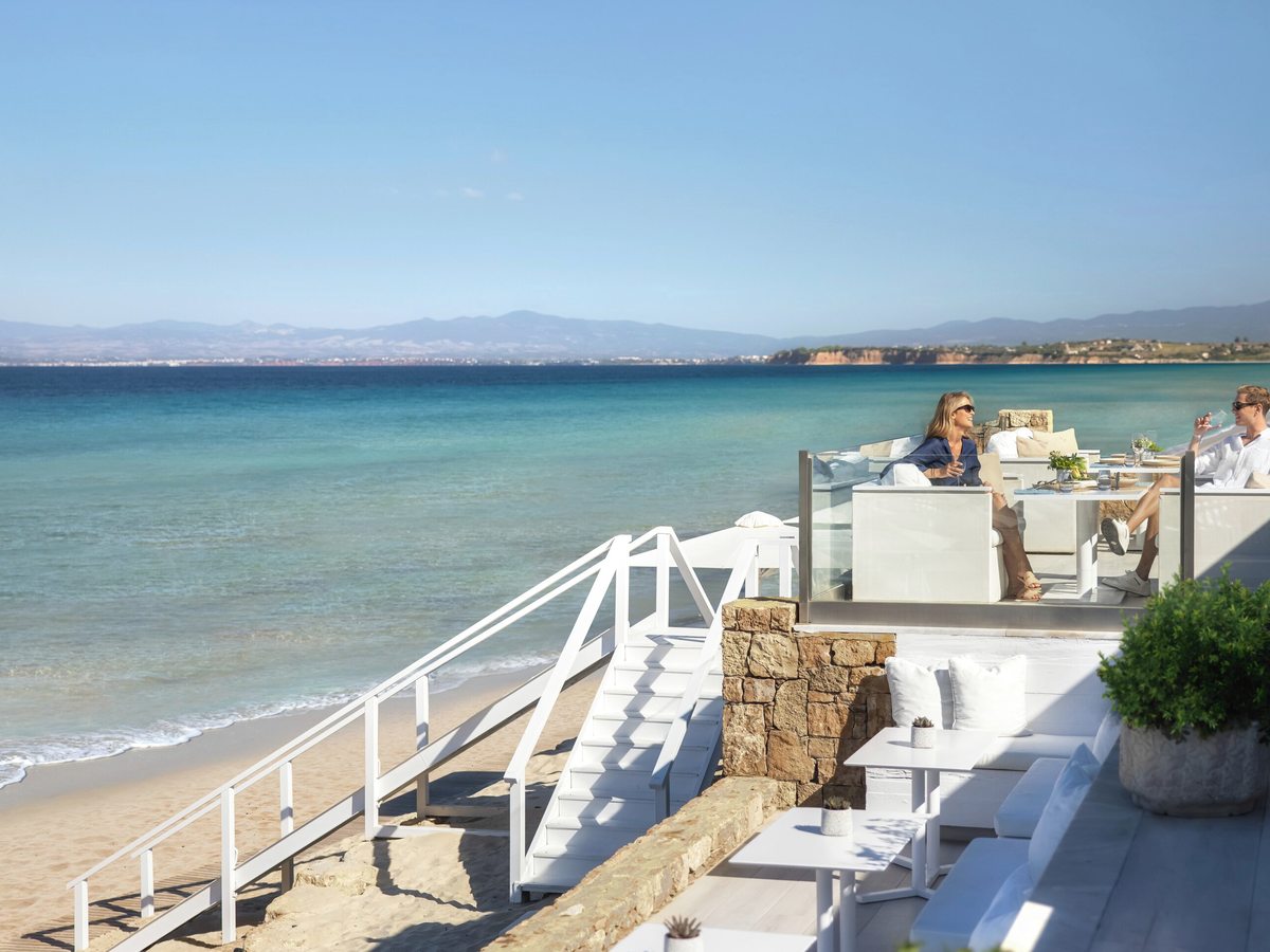 White-painted restaurant terrace above a sandy beach, with steps down to the sand, cushioned seating and two guests at a table, the bay and distant mountains behind.
