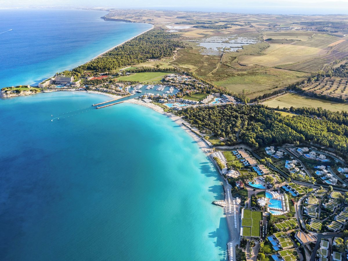 Aerial view of a wooded cape with a curving turquoise bay, a marina full of boats, low white hotel buildings among pine forest, and farmland stretching inland.