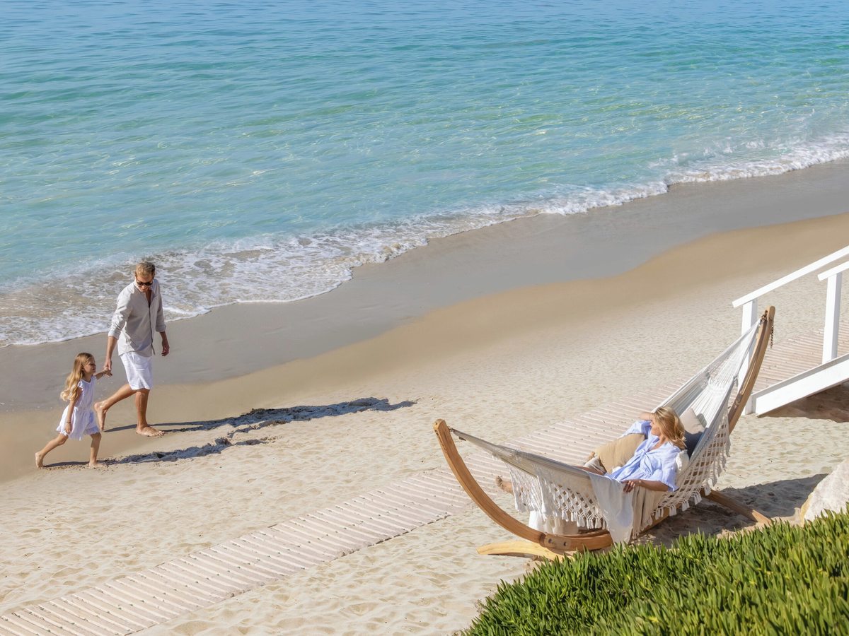 Wooden boardwalk crossing soft sand towards the water's edge, with a hammock on a wooden stand, a woman resting in it and a man and a young girl walking along the shoreline.