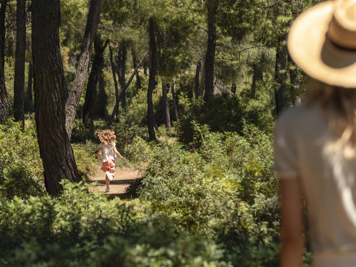 A young child running ahead along a bare earth trail through tall pine trees, with a woman in a straw sunhat out of focus in the foreground.