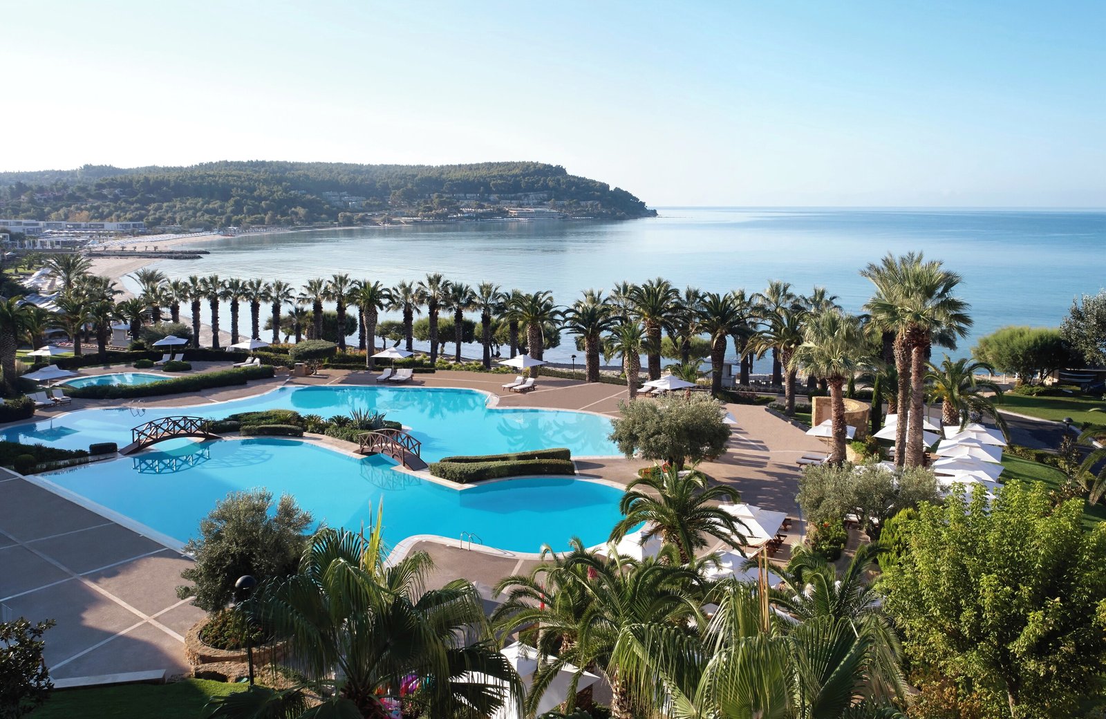 The lagoon-shaped main pool at Sani Beach, crossed by two small bridges and edged with palms, loungers and white parasols, with the white-sand beach and the wooded bay of Cape Sani behind.