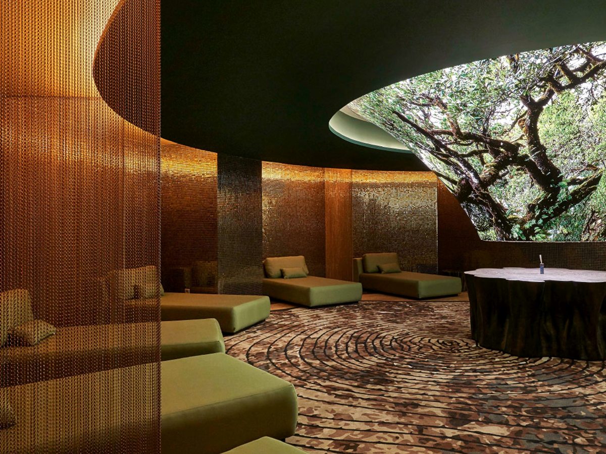 A curved relaxation lounge with green cushioned loungers, gold chainmail curtains and a large oval opening onto an old laurel tree.