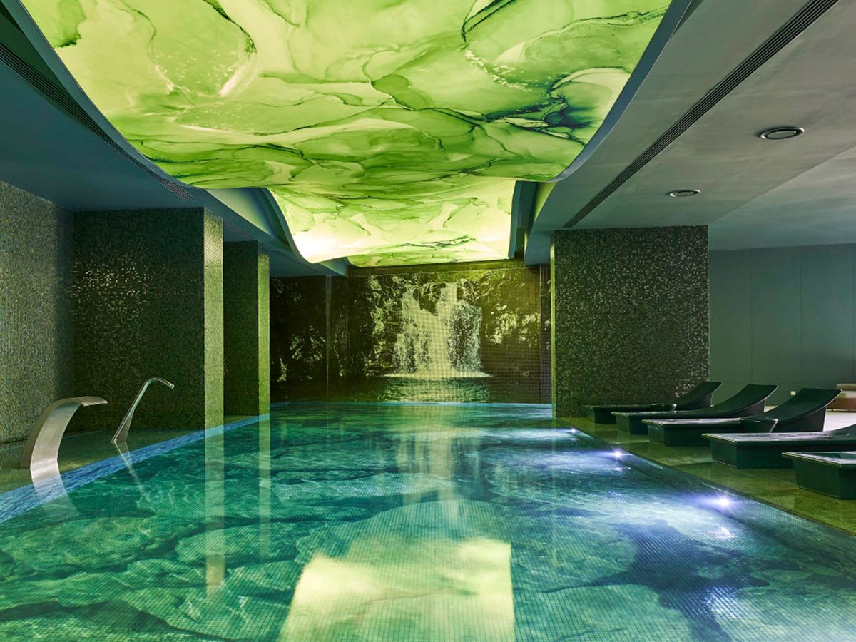 A long indoor pool under a backlit green marbled ceiling, with a waterfall mural at the far end and dark loungers along one side.