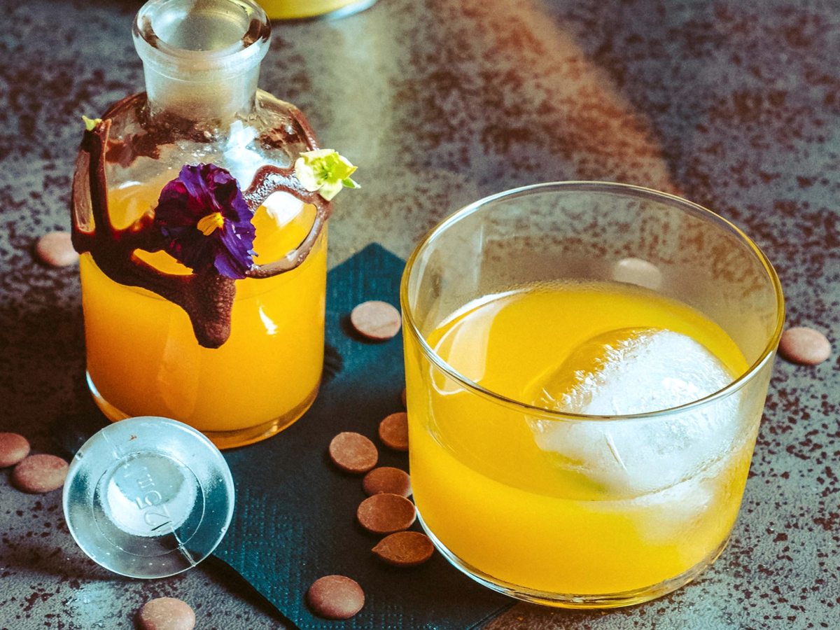 A tumbler of golden cocktail over a large ice cube next to a glass carafe of the same drink garnished with an edible flower.