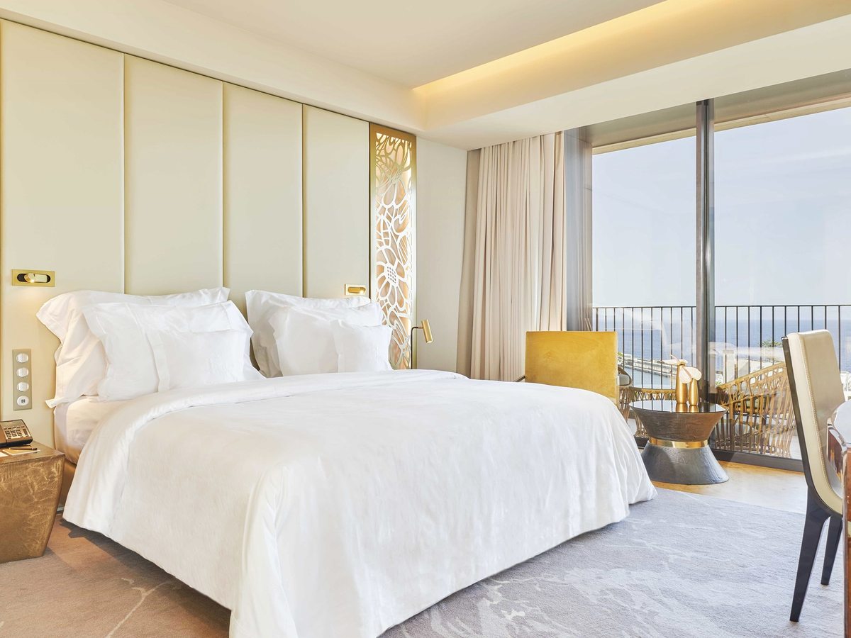 A cream and gold bedroom with a king bed, a padded headboard wall inset with a gold filigree panel and glass doors onto a balcony above the sea.